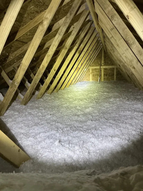 New-build attic with blown-in fiberglass insulation and roof trusses lit by an inspector's flashlight